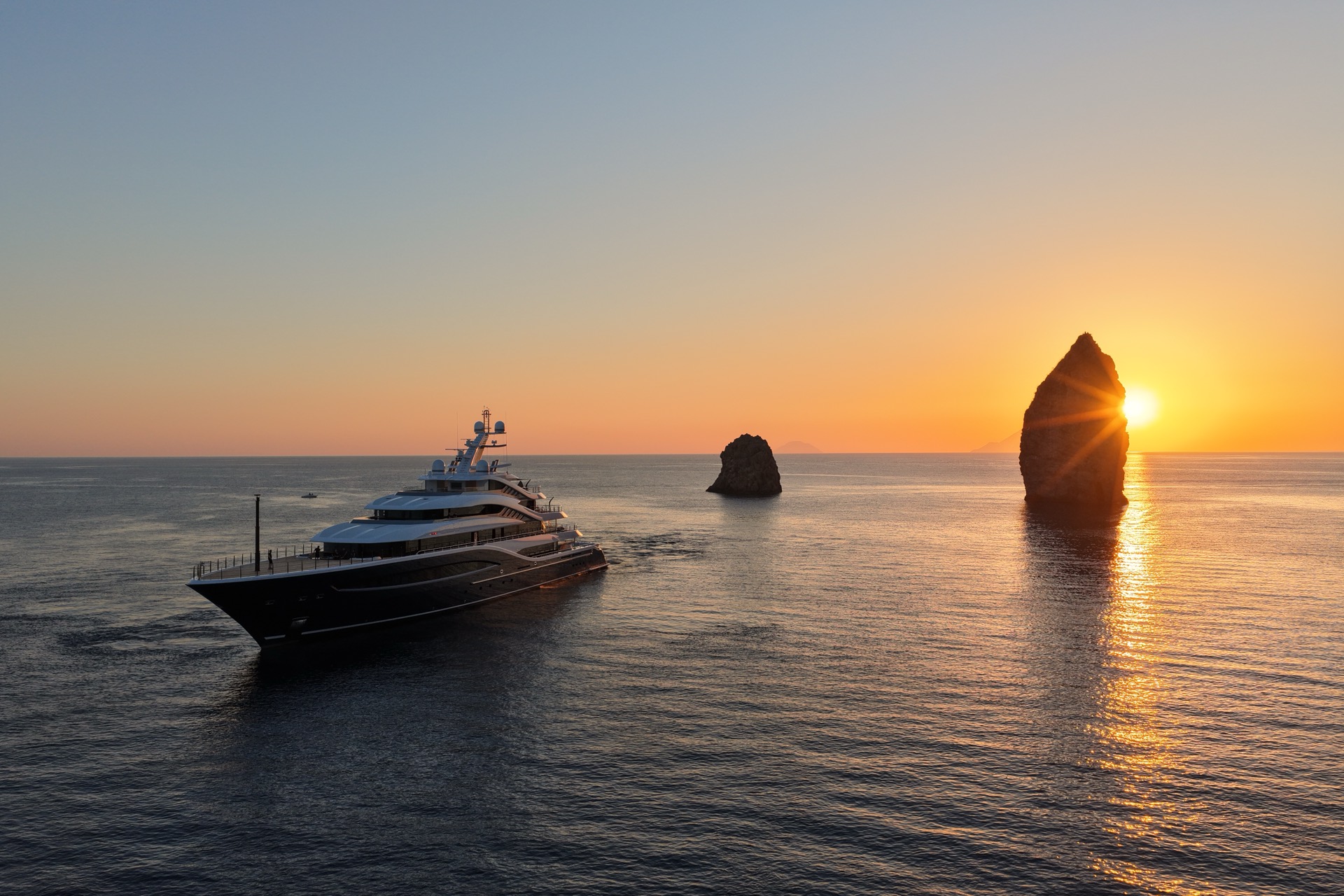 A superyacht at sunset beside a tall sea stack, with the sun's reflection glittering across the water