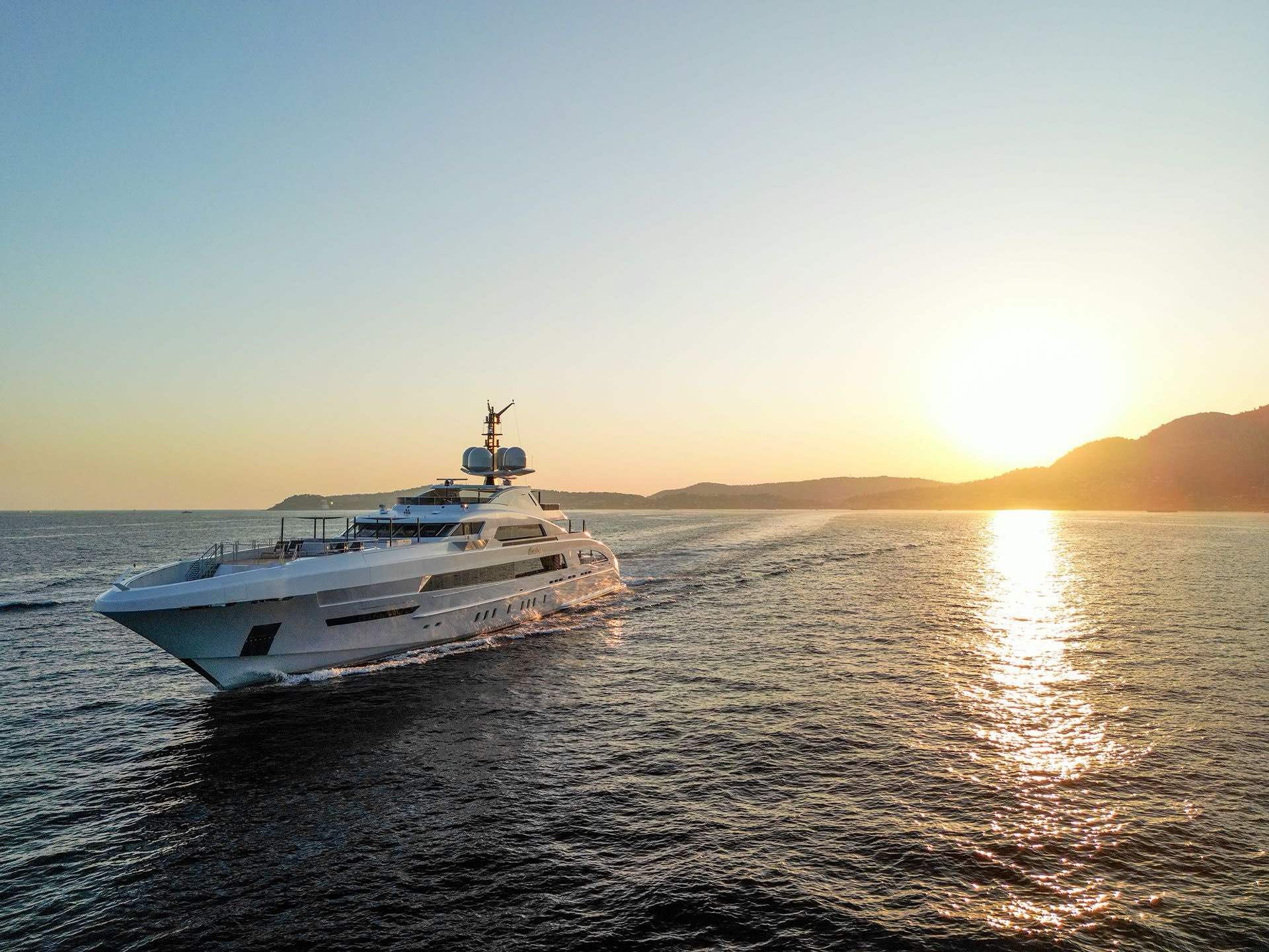 A superyacht under way at sunset, with the coastline behind