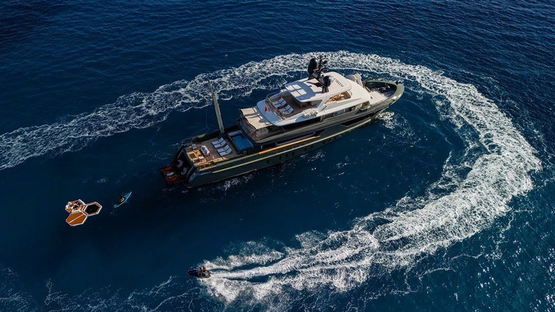 A motor yacht under way seen from above, with a tender circling her and a sea platform alongside