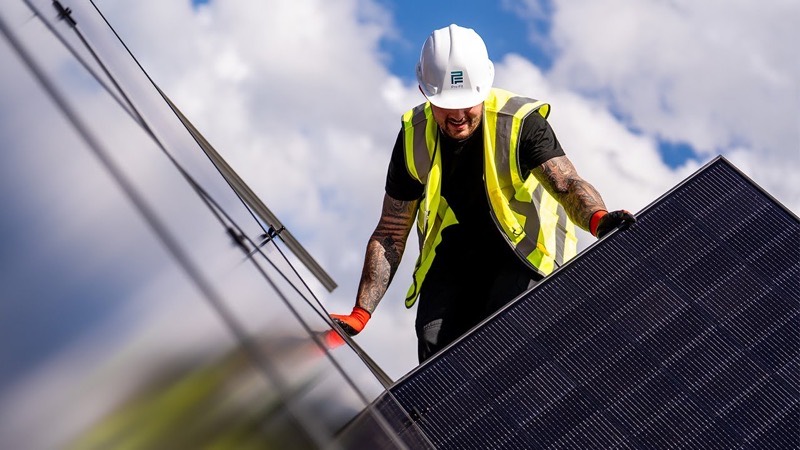 An installer in a hard hat fitting a solar panel against a bright sky